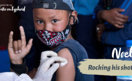 Boy wearing mask making rock n roll gesture as he receives a vaccine. text: "Noel: Rocking his shot"