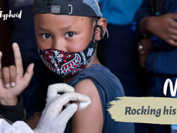 Boy wearing mask making rock n roll gesture as he receives a vaccine. text: "Noel: Rocking his shot"