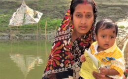 Woman holds a young child with a vaccination card, with water in the background