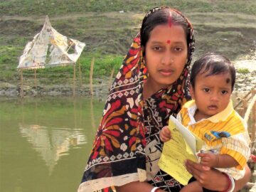 Woman holds a young child with a vaccination card, with water in the background