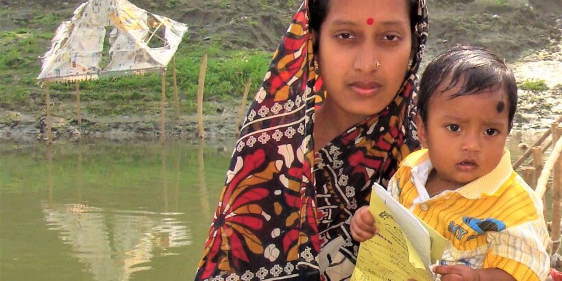 Woman holds a young child with a vaccination card, with water in the background