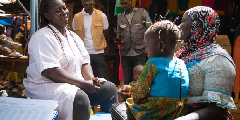 A health care worker in a medical uniform speaks to a woman holding her child during a typhoid conjugate vaccine campaign in Burkina Faso.