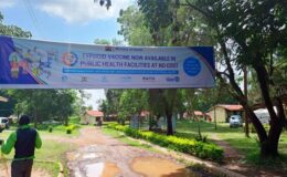 A large banner hangs over a street in a town on a sunny day. The banner advertises the upcoming typhoid conjugate vaccine introduction campaign in Kenya.