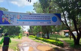 A large banner hangs over a street in a town on a sunny day. The banner advertises the upcoming typhoid conjugate vaccine introduction campaign in Kenya.