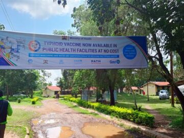 A large banner hangs over a street in a town on a sunny day. The banner advertises the upcoming typhoid conjugate vaccine introduction campaign in Kenya.