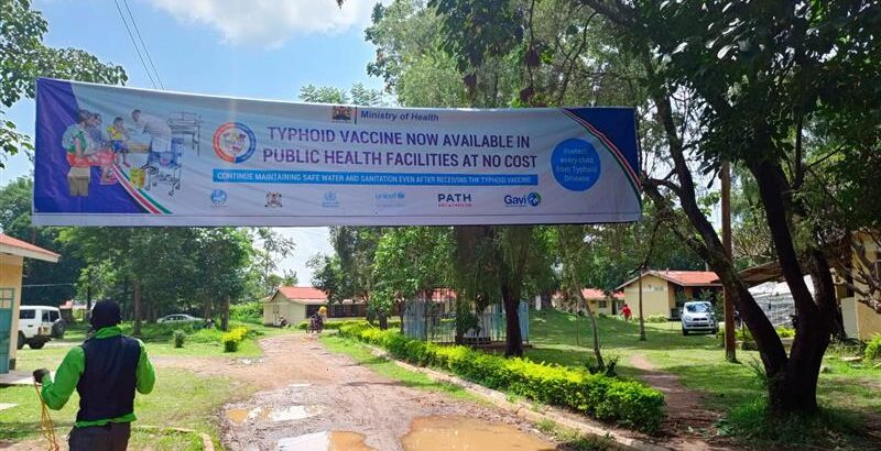 A large banner hangs over a street in a town on a sunny day. The banner advertises the upcoming typhoid conjugate vaccine introduction campaign in Kenya.