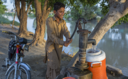 A young boy uses a pump to pump water into an orange and white container on the banks of a river.