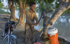 A young boy uses a pump to pump water into an orange and white container on the banks of a river.