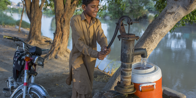 A young boy uses a pump to pump water into an orange and white container on the banks of a river.