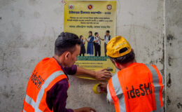 Two campaign workers wearing orange vests put up a sign with information about the typhoid conjugate vaccine in Bangladesh.