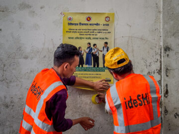Two campaign workers wearing orange vests put up a sign with information about the typhoid conjugate vaccine in Bangladesh.