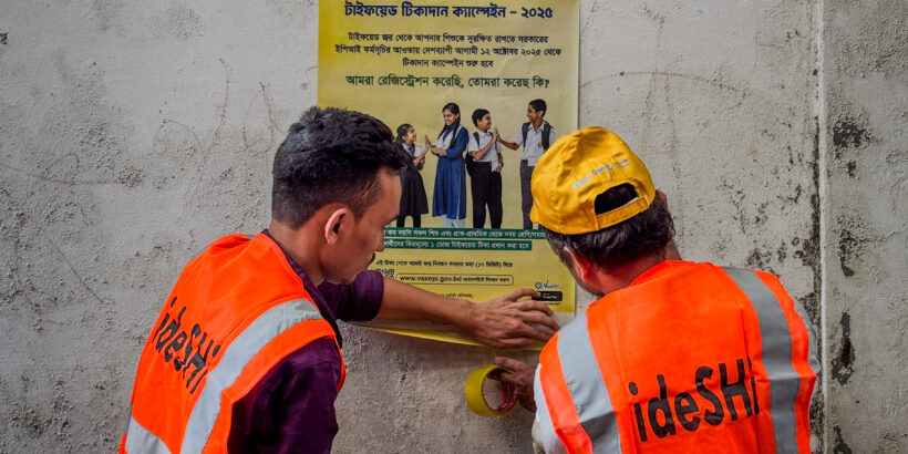 Two campaign workers wearing orange vests put up a sign with information about the typhoid conjugate vaccine in Bangladesh.